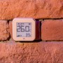 Beiger Shelly Temperatursensor Plus H&T G3 an einer Ziegelwand. Zeigt Temperatur und Luftfeuchtigkeit.