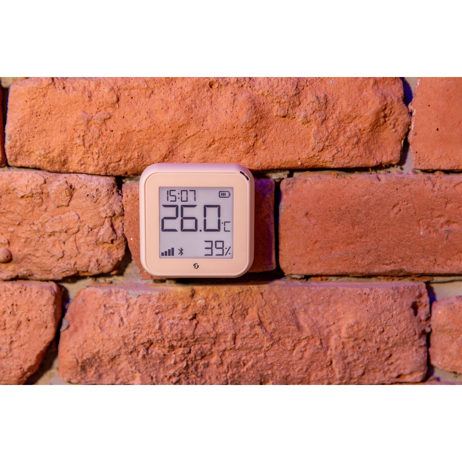 Beiger Shelly Temperatursensor Plus H&T G3 an einer Ziegelwand. Zeigt Temperatur und Luftfeuchtigkeit.