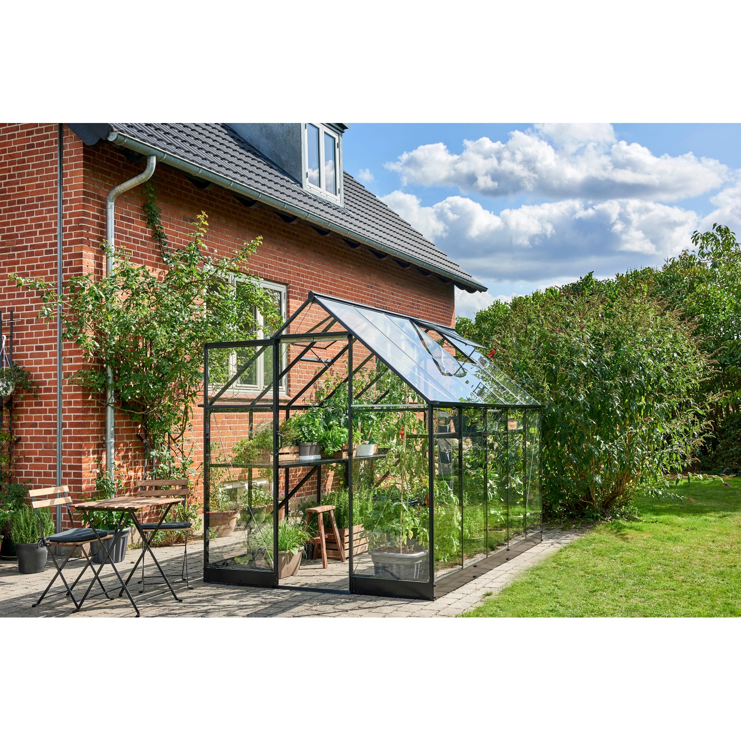 Schwarzes Halls Gewächshaus Qube 610 mit Pflanzen und Sitzgruppe im Garten.
