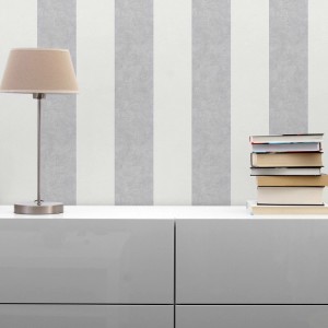 Tadessi Vliestapete mit cremefarbenen und weißen Streifen an einer Wand, daneben Lampe und Bücher. Dekorative Tapete.