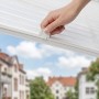 Weiße Plissee Klemmfix Faltrollo am Fenster, eine Hand verstellt den blickdichten Sonnenschutz. Ohne Bohren, für Fenster und Türen.