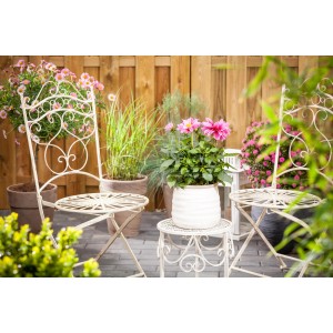 Rosa Dahlie 'Dahlietta Emily' im Topf auf Gartentisch, umgeben von Gartenmöbeln und Pflanzen.