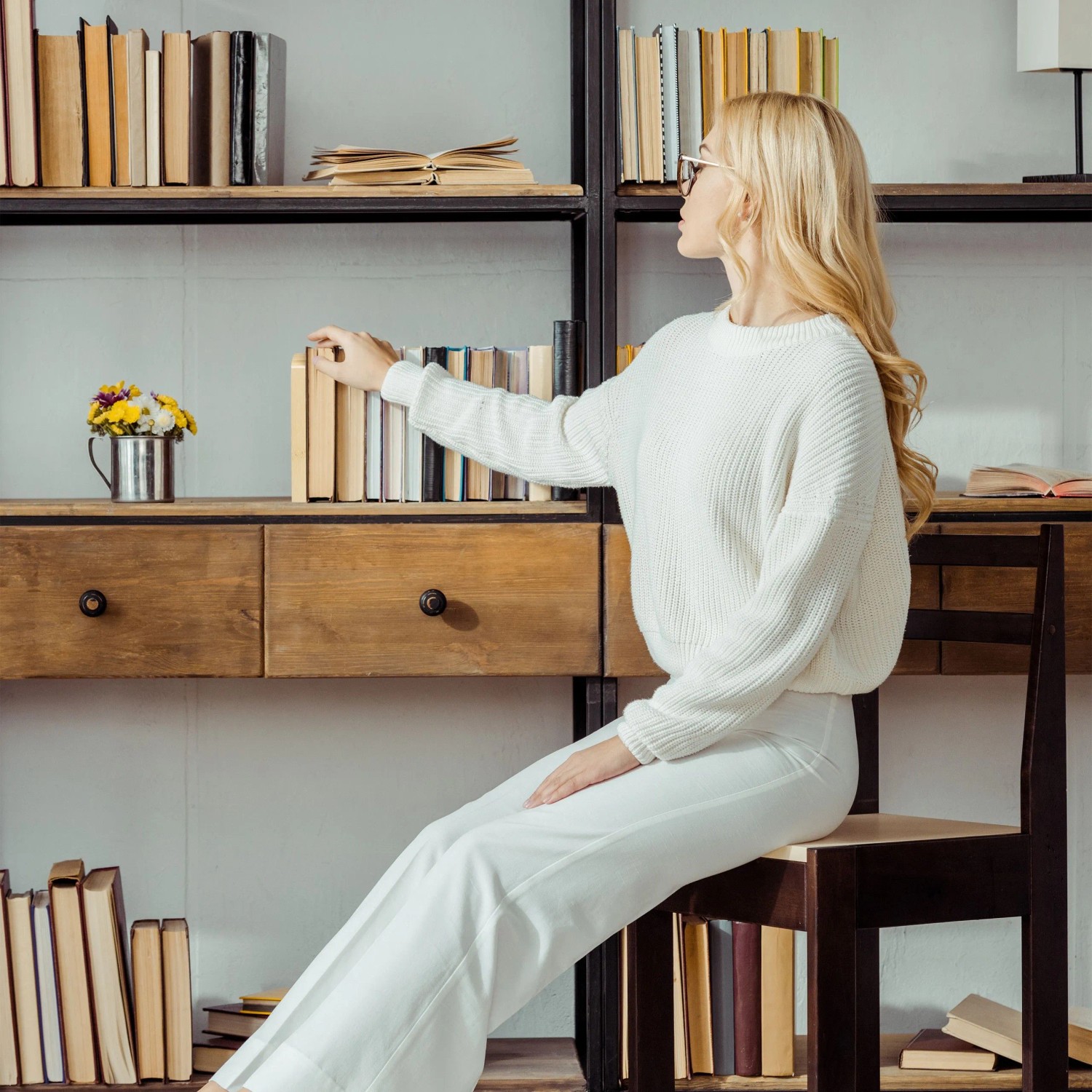 Blonde Frau in weißem Pullover wählt ein Buch aus einem Holzregal. Perfekt für Buchstützen aus Bambus und Edelstahl im Büro.