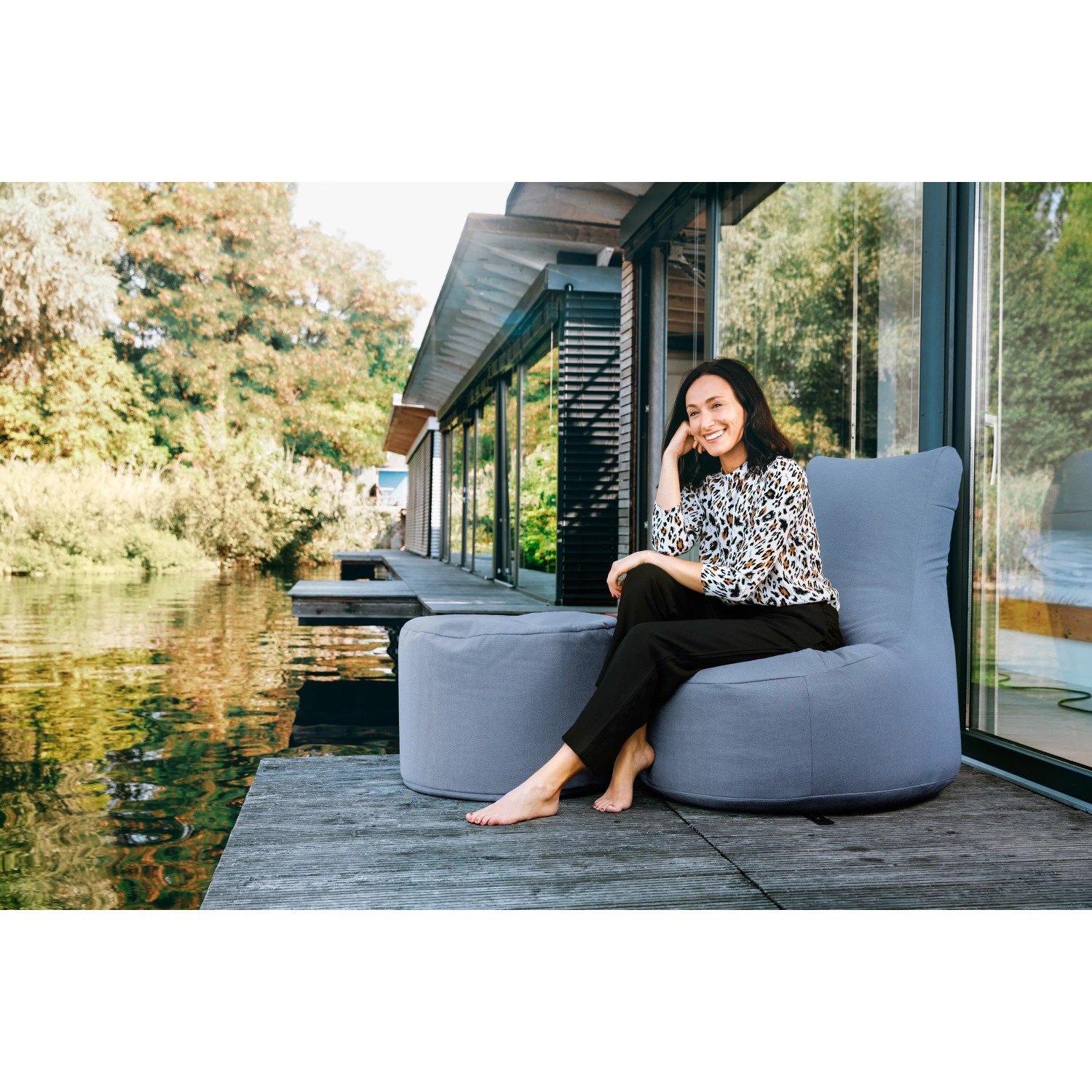 Frau sitzt auf blauem Sitting Point Hocker Outside (120 l) am Wasser. Bequemer Sitzsack für Outdoor & Indoor.