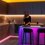 Moderne Küche mit Frau, beleuchtet von Philips Smart GU10 RGB LED-Licht. Farbige Akzente unter der Theke, warmweißes Arbeitslicht.