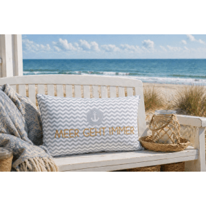 Dekokissen mit grau-weißem Chevron-Muster, Anker und "Meer geht immer" Schriftzug auf einer Holzbank am Strand.