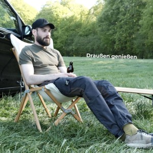 Mann sitzt entspannt in einem Holz Faltsessel mit hellem Bezug im Grünen, neben einem Auto. Ideal für Camping.