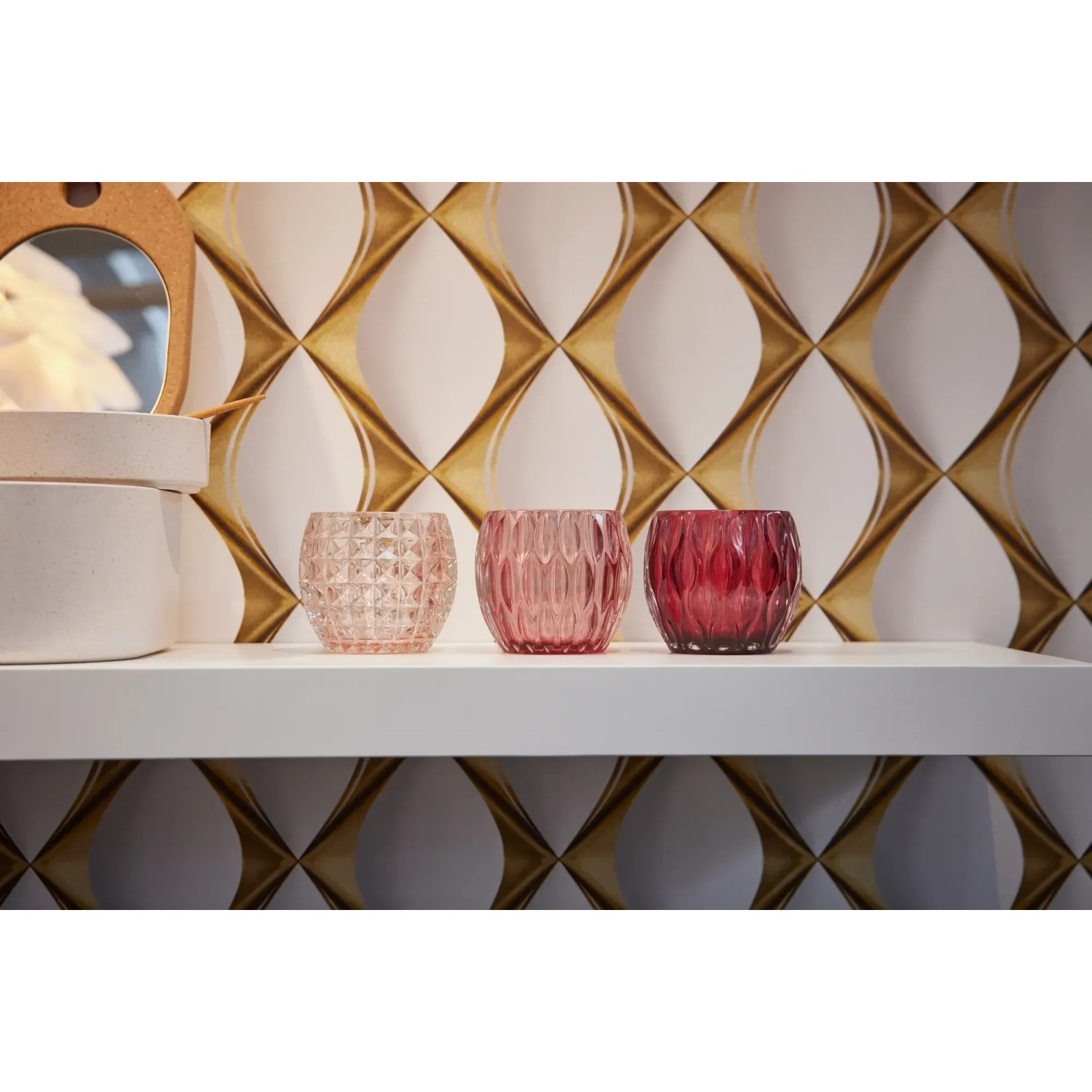 Nahaufnahme von drei Teelichthaltern in Rosa- und Rottönen vor einer goldenen Vliestapete mit 3D-Design.