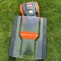 YARD FORCE Rasenmähroboter Compact 300RBS und Mähroboter-Garage auf grünem Rasen. Orange-schwarzer Mähroboter unter Schutzdach.