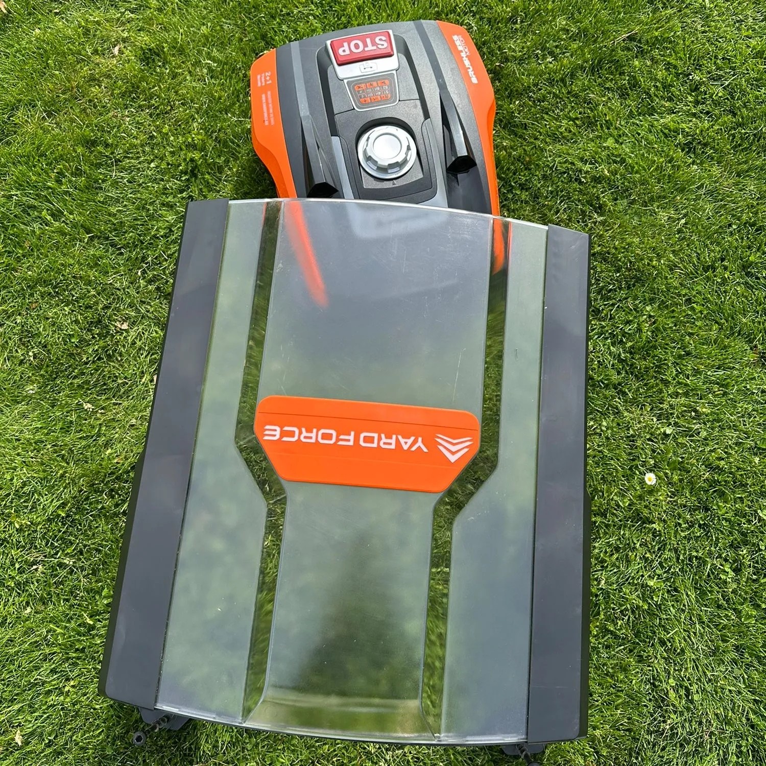 YARD FORCE Rasenmähroboter Compact 300RBS und Mähroboter-Garage auf grünem Rasen. Orange-schwarzer Mähroboter unter Schutzdach.
