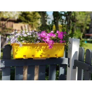 Gelber GreenLife Blumenkasten mit lila und pinkfarbenen Blumen auf einem Zaun.