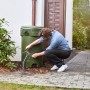 Mann repariert mit transparentem Tesa Reparaturband ein grünes Regenfass im Garten.