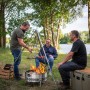 Petromax Hänge-Rost am Dreibein über Feuerstelle mit grillenden Personen im Freien.