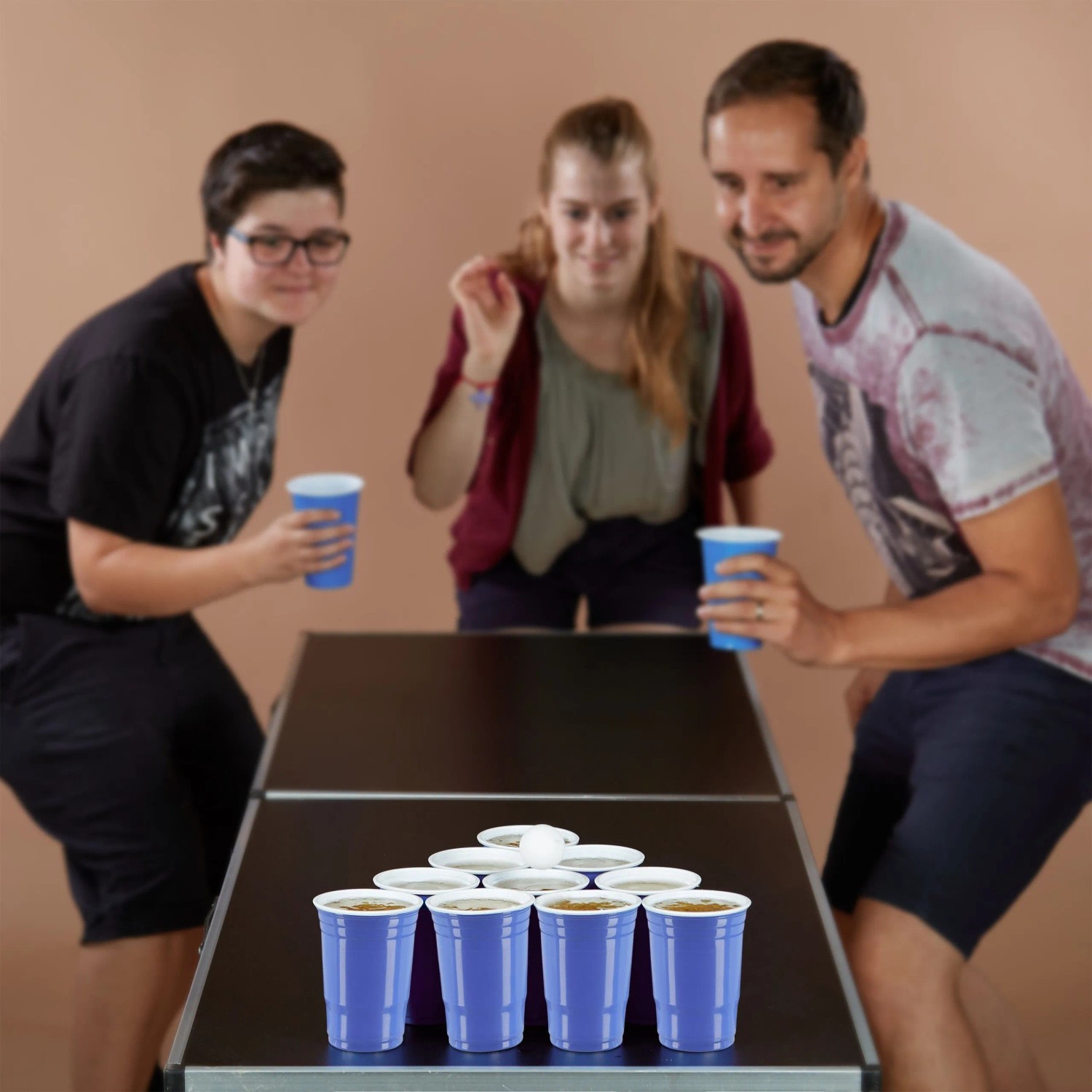 Drei Personen spielen Bierpong mit blauen Kunststoffbechern und einem Ball auf einem Tisch. Party-Set für gesellige Abende.