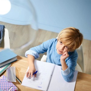 Blonder Junge sitzt am Schreibtisch, macht Hausaufgaben und blickt nachdenklich nach oben, beleuchtet von einem Deckenstrahler.