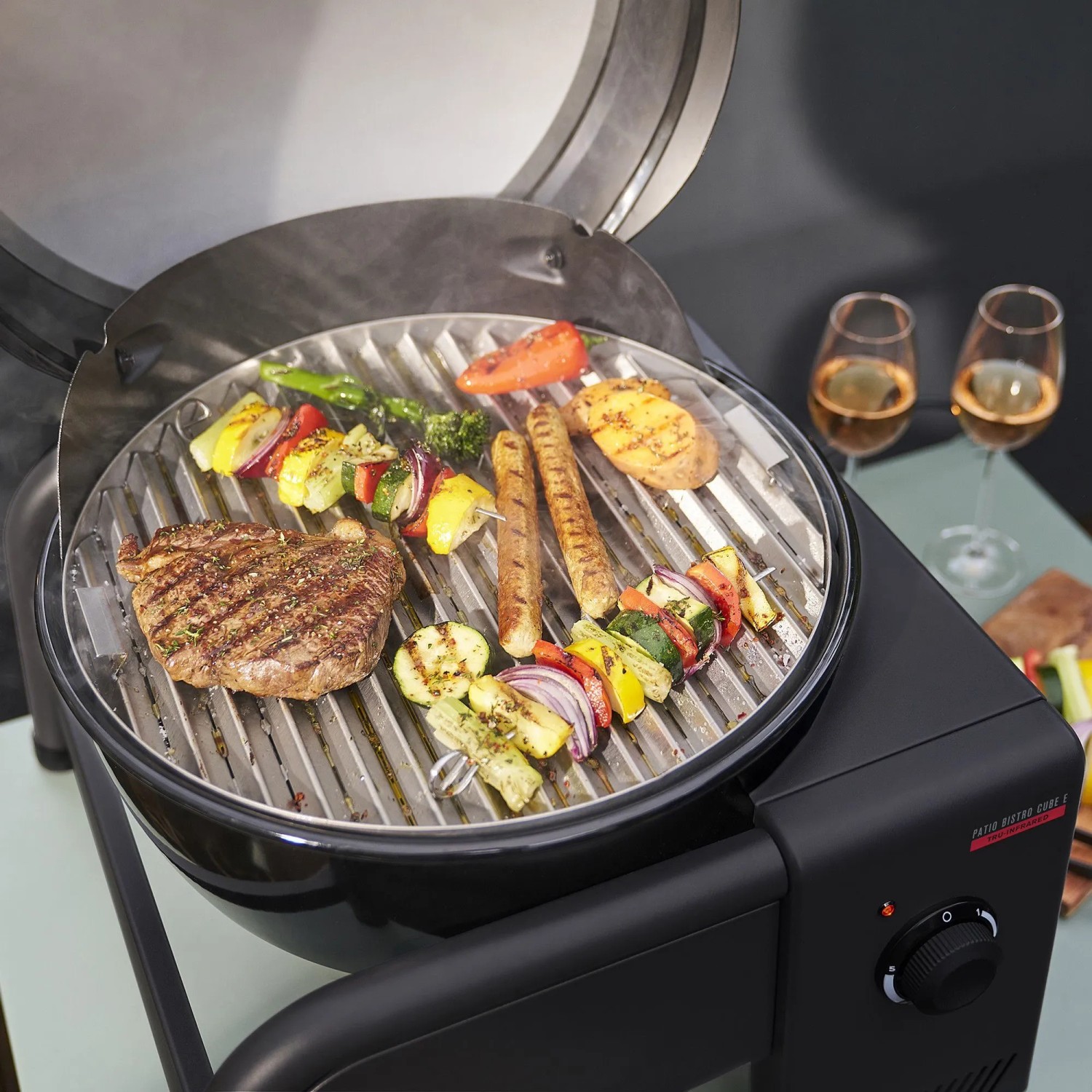 Schwarzer Char-Broil Elektrogrill Patio Bistro Cube E mit Grillgut und zwei Gläsern Wein.