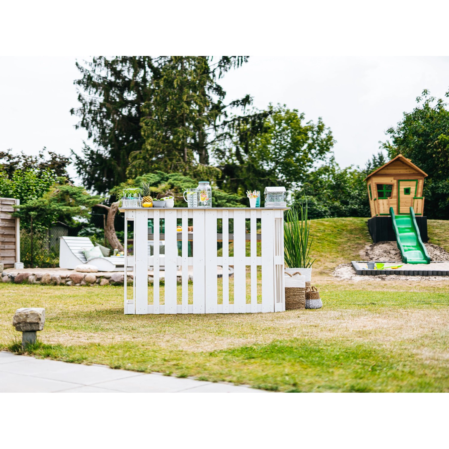 Axi Spielhaus Max mit Rutsche und Leiter im Garten, im Hintergrund eine DIY-Bar aus Paletten.