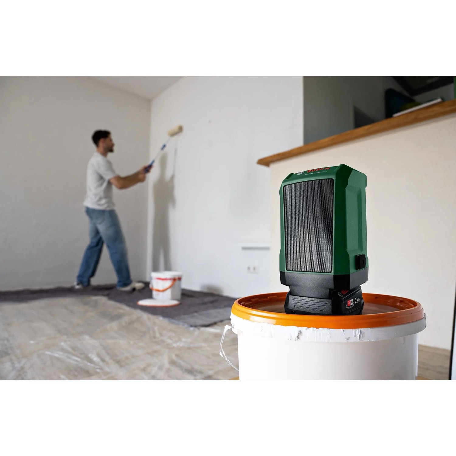 Grüner Bosch Akku-Bluetooth-Box UniversalSpeaker 18V-1 auf Eimer, Mann streicht Wand.