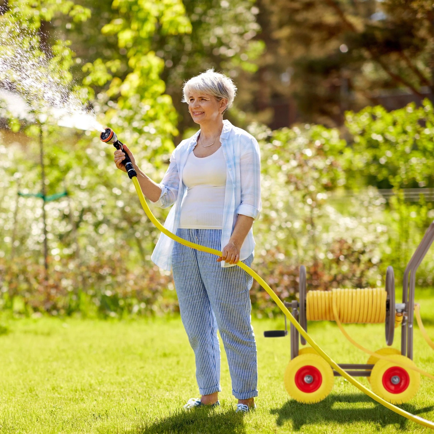 Frau bewässert Garten mit gelbem Schlauch. Mobiler Gartenschlauchwagen aus Stahl mit 4 Rädern für einfache Bewässerung.