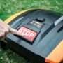 Hand drückt roten STOP-Knopf auf schwarzem Mähroboter im grünen Rasen.