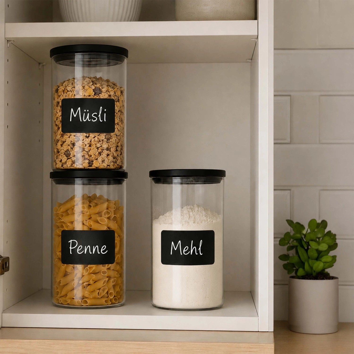 3er Set transparente Vorratsgläser mit schwarzen Deckeln und Etiketten, gefüllt mit Müsli, Nudeln und Mehl im Küchenschrank.