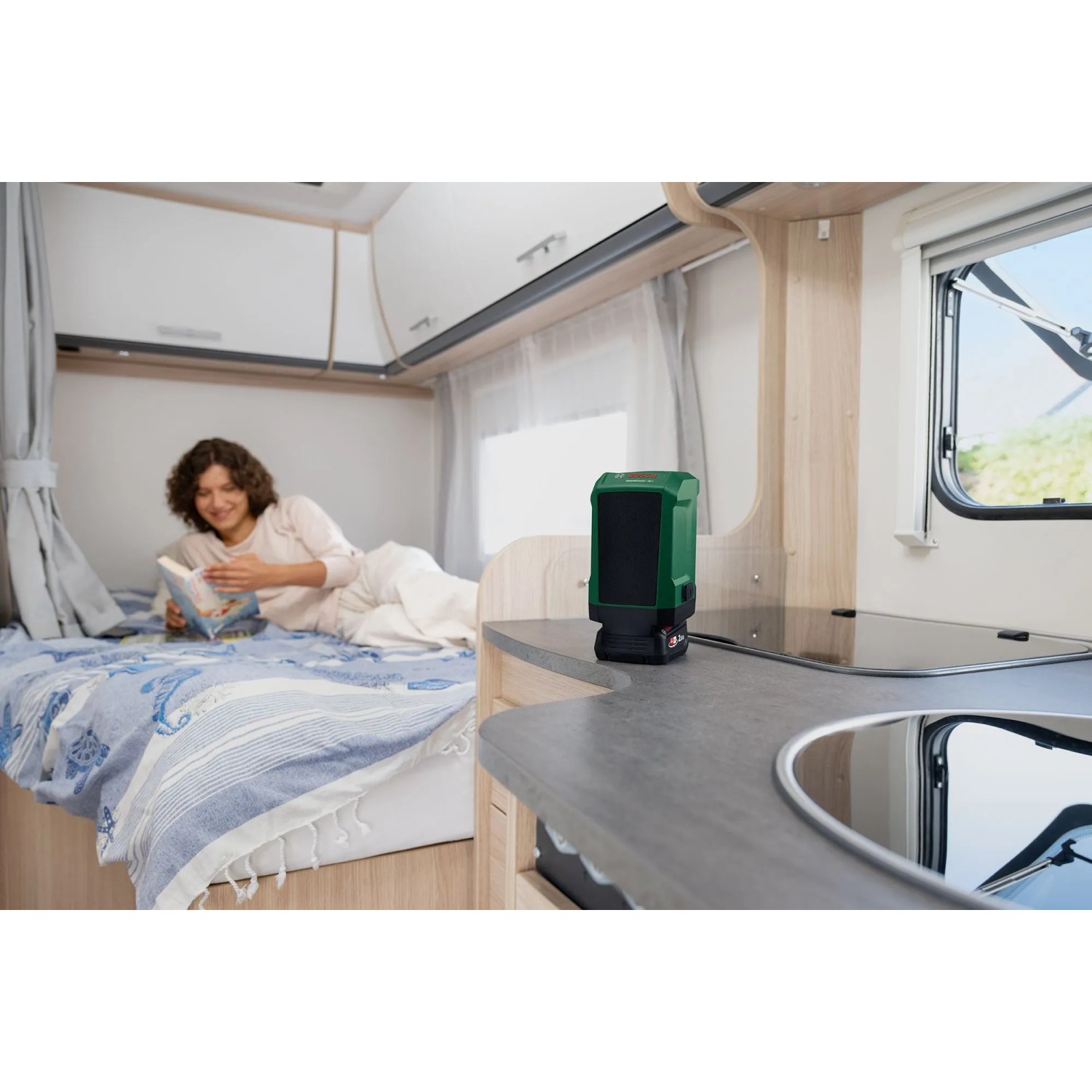 Dunkelgrüner Bosch UniversalSpeaker 18V-1 Akku-Bluetooth-Lautsprecher auf Arbeitsplatte im Wohnmobil. Frau liest im Bett.