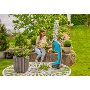 Gardena Akku-Gras- und Strauchschere PowerCut 20/18V Set auf Gartentisch, im Hintergrund Frau im Garten.