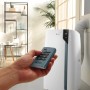 DeLonghi Klimagerät mit Fernbedienung in der Hand vor Fenster und Regal.