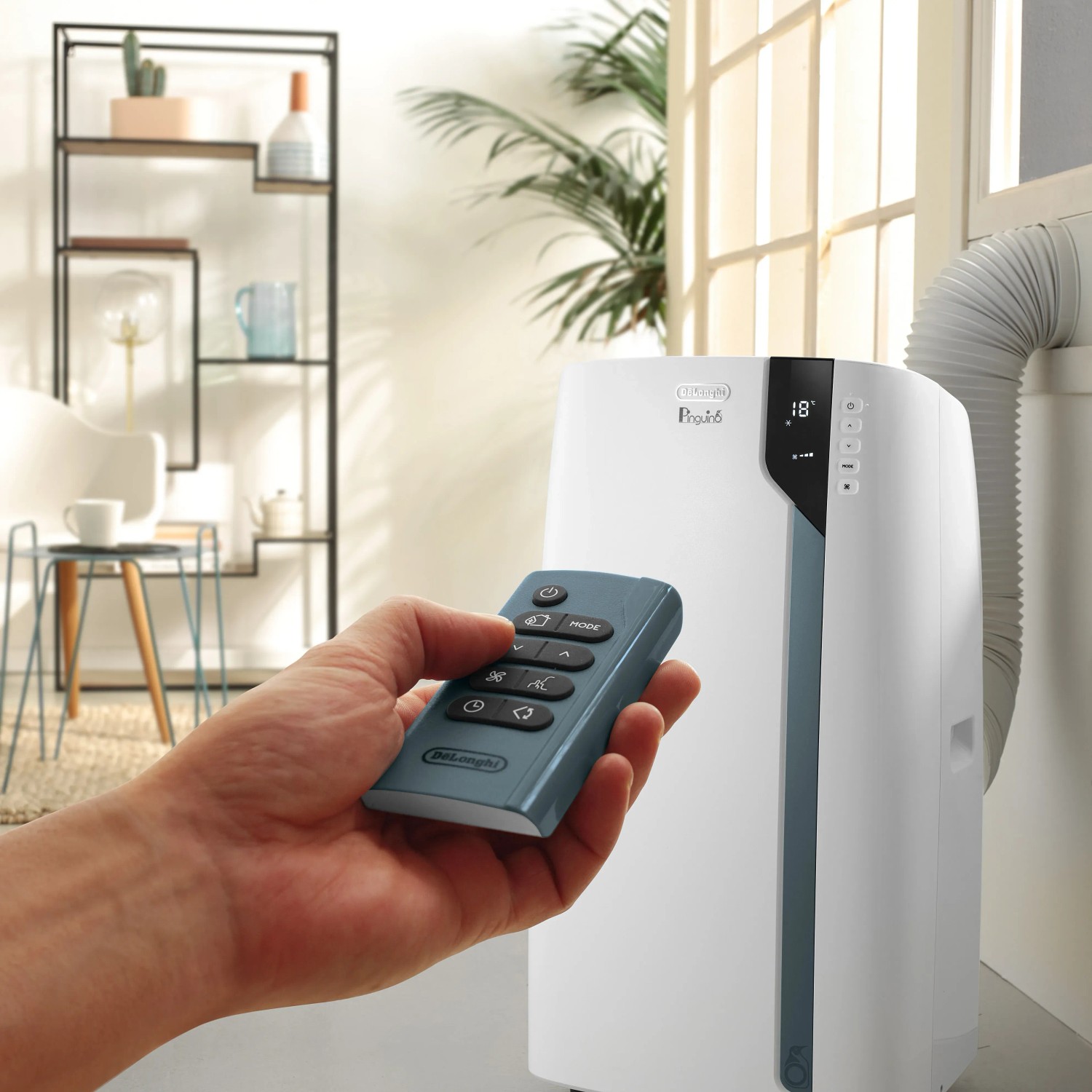 DeLonghi Klimagerät mit Fernbedienung in der Hand vor Fenster und Regal.