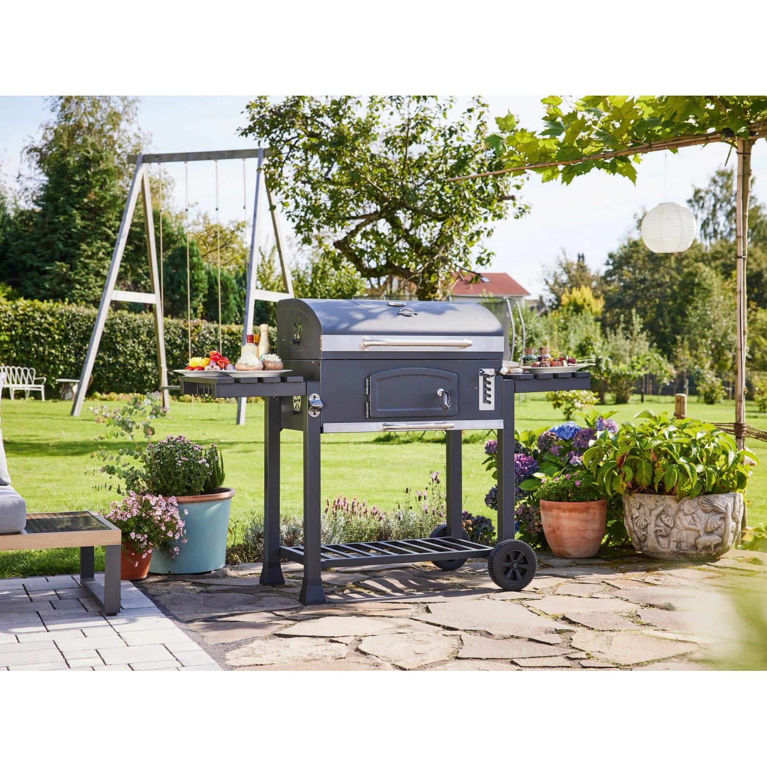 Jamestown Holzkohlegrillwagen Jaxon XL im Garten mit Zubehör auf Seitentischen.
