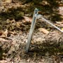 Silberfarbener AMANKA V-Zelthering aus verzinktem Stahl im Waldboden mit weißer Spannleine. Ideal für Camping.