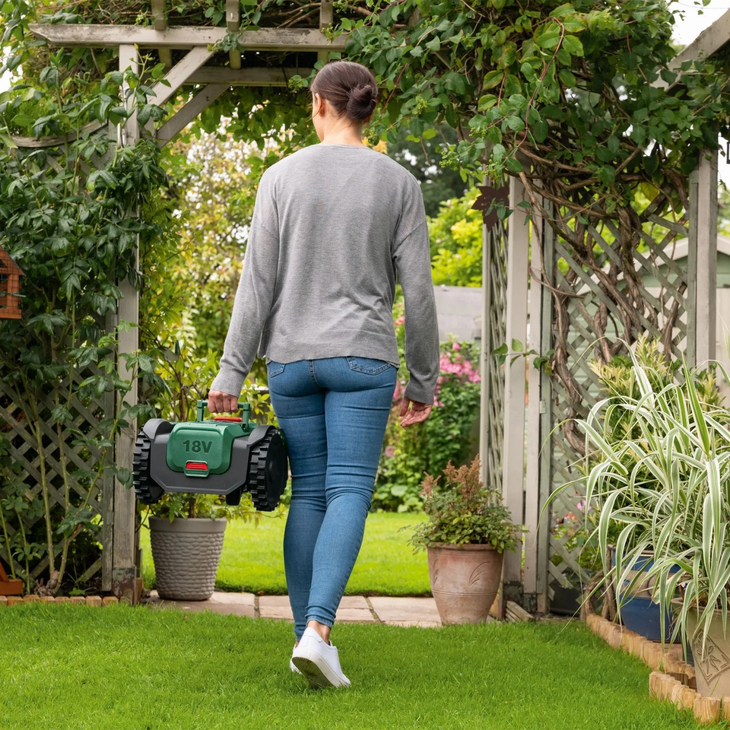 Frau trägt Bosch Mähroboter VISIMOW18V-100 durch den Garten.