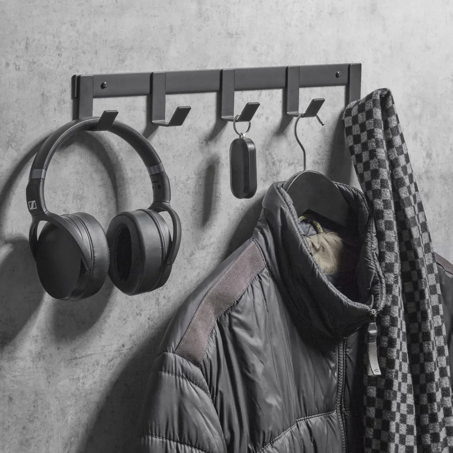 Schwarze SOTECH Blackline ALBA Hakenleiste mit 4 Haken an grauer Wand, behängt mit Kopfhörern, Schlüssel, Jacke. Garderobenleiste.
