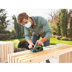 Mann hobelt Holzbank mit Bosch Elektrohobel PHO 20 im Garten. Grüner Hobel, Schutzbrille, Maske für präzise Holzbearbeitung.