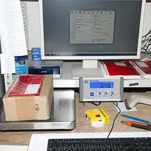 PCE Instruments Plattformwaage mit LC Display auf einem Schreibtisch mit PC und Paket.