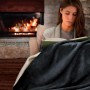 Frau liest gemütlich unter einer anthrazitfarbenen Beautissu Alina Kuscheldecke mit weißer Lammfelloptik vor Kaminfeuer.