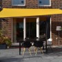 Sonnengelbes Vierecksonnensegel 300x400 cm mit Regenschutz über einer Terrasse mit Tisch und Stühlen.
