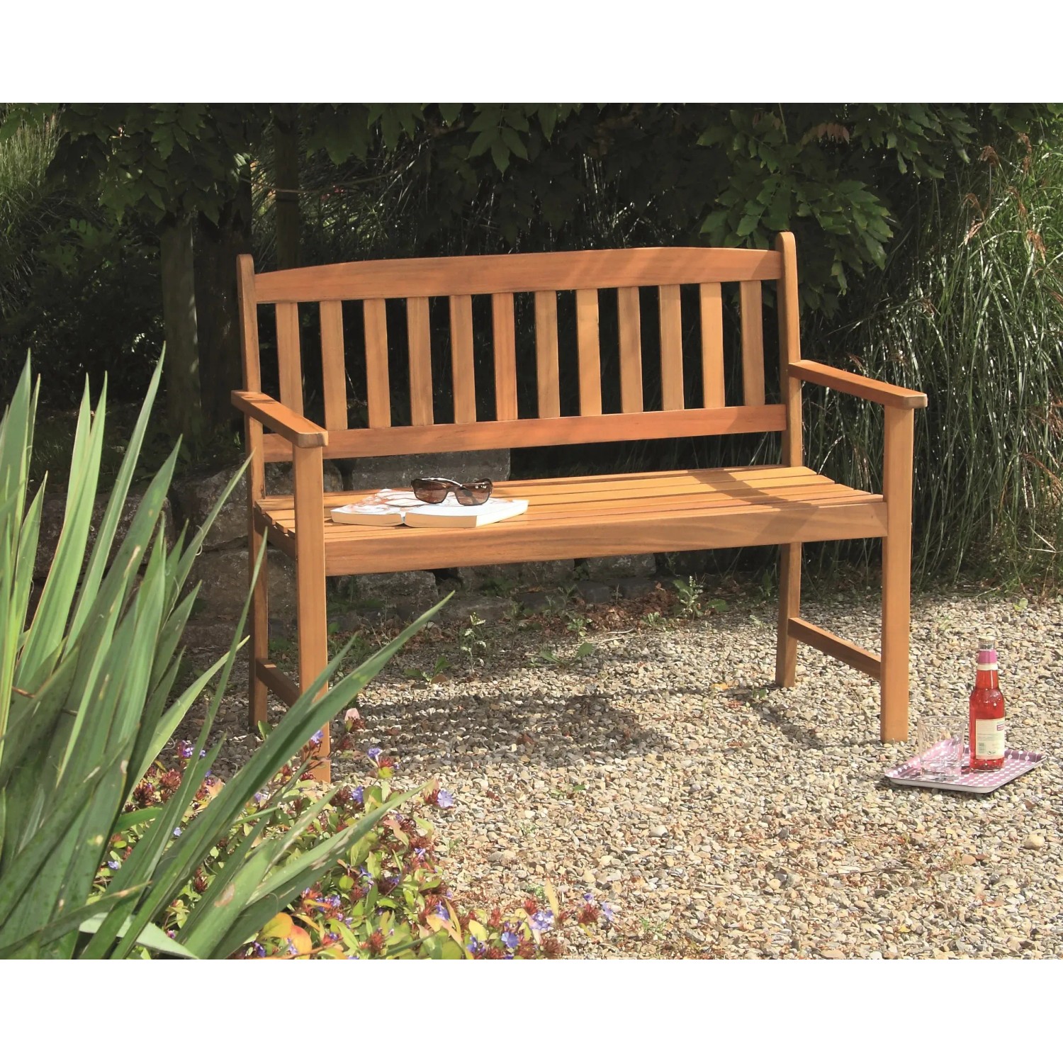 Acamp Gartenbank Woody 2-Sitzer aus Teakholz (FSC®) im Garten, mit Buch und Getränk dekoriert.