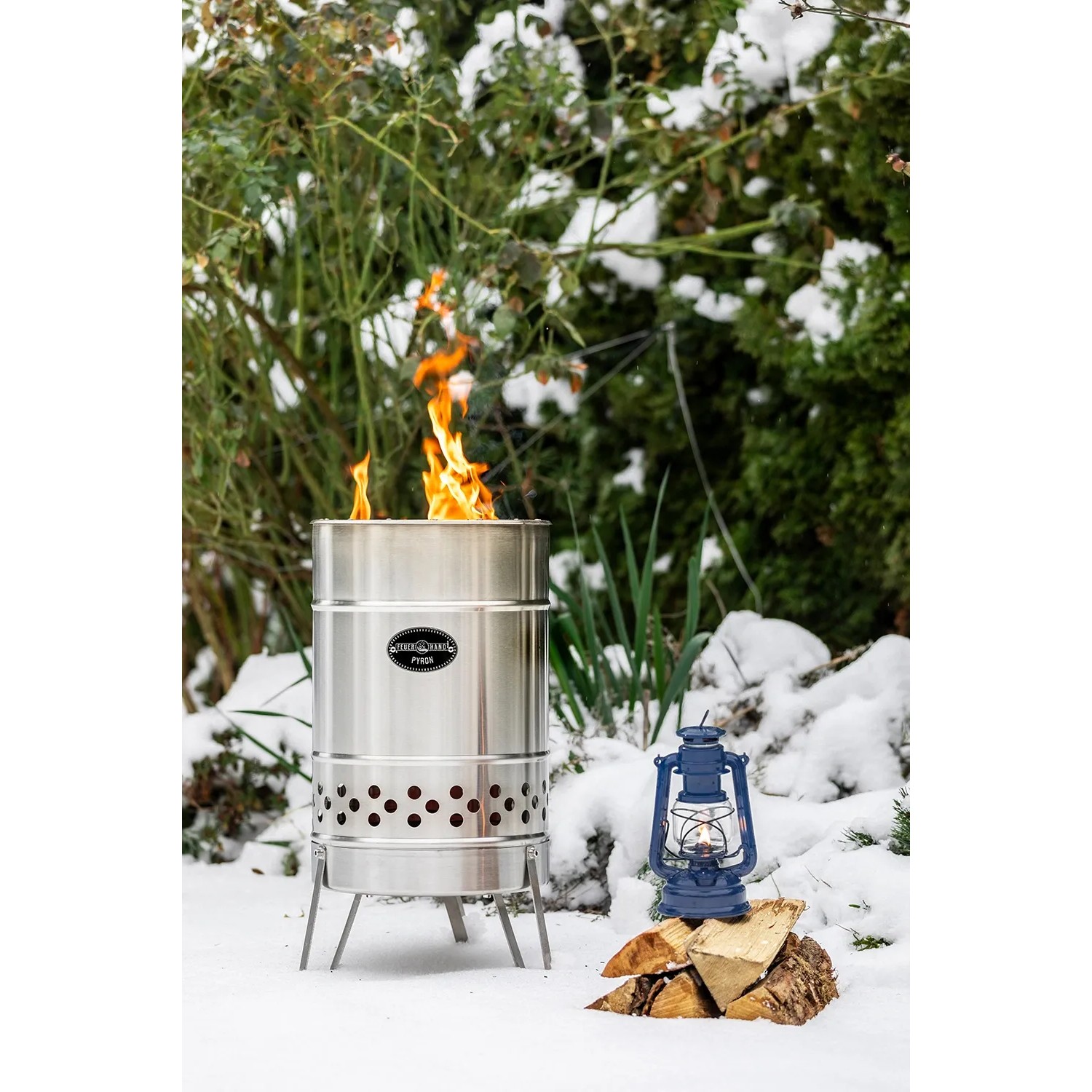 Feuerhand Pyron Feuertonne aus Edelstahl (25 l) mit Flammen und blauer Laterne im Garten.