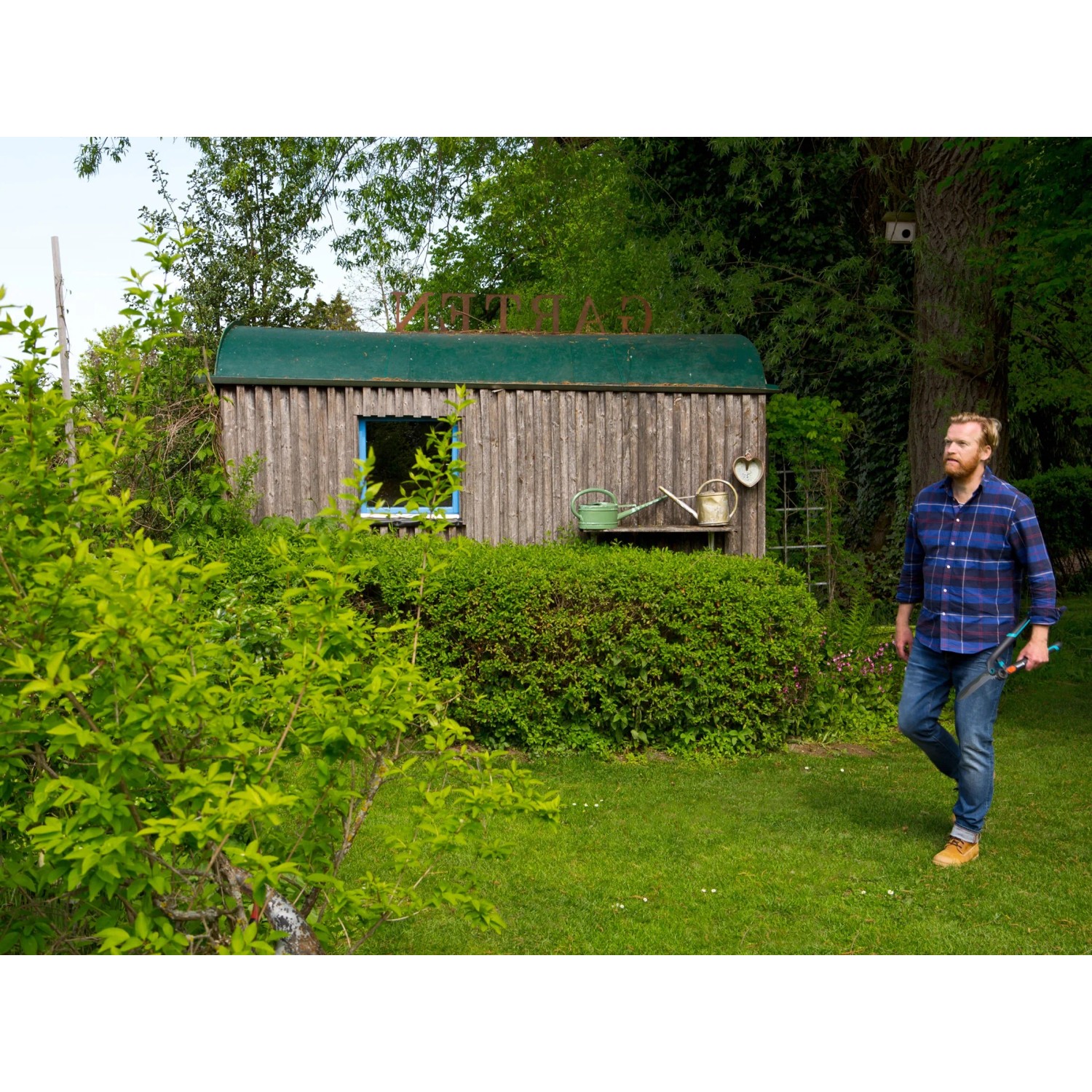 Mann mit Gardena Heckenschere 2in1 EnergyCut im Garten vor Holzhütte.