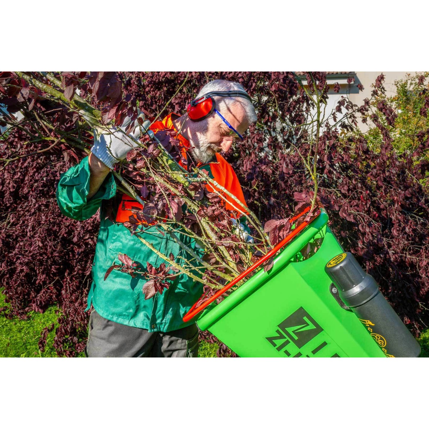 Mann bedient Zipper ZI-HAEK4100 Elektro-Häcksler mit Ästen vor Garten.