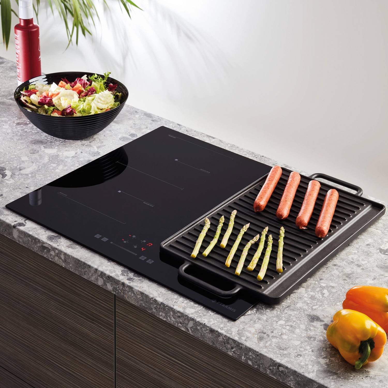 Schwarzes Pyramis Induktionskochfeld PHI63432FMB mit Grillplatte, Würstchen und Spargel.