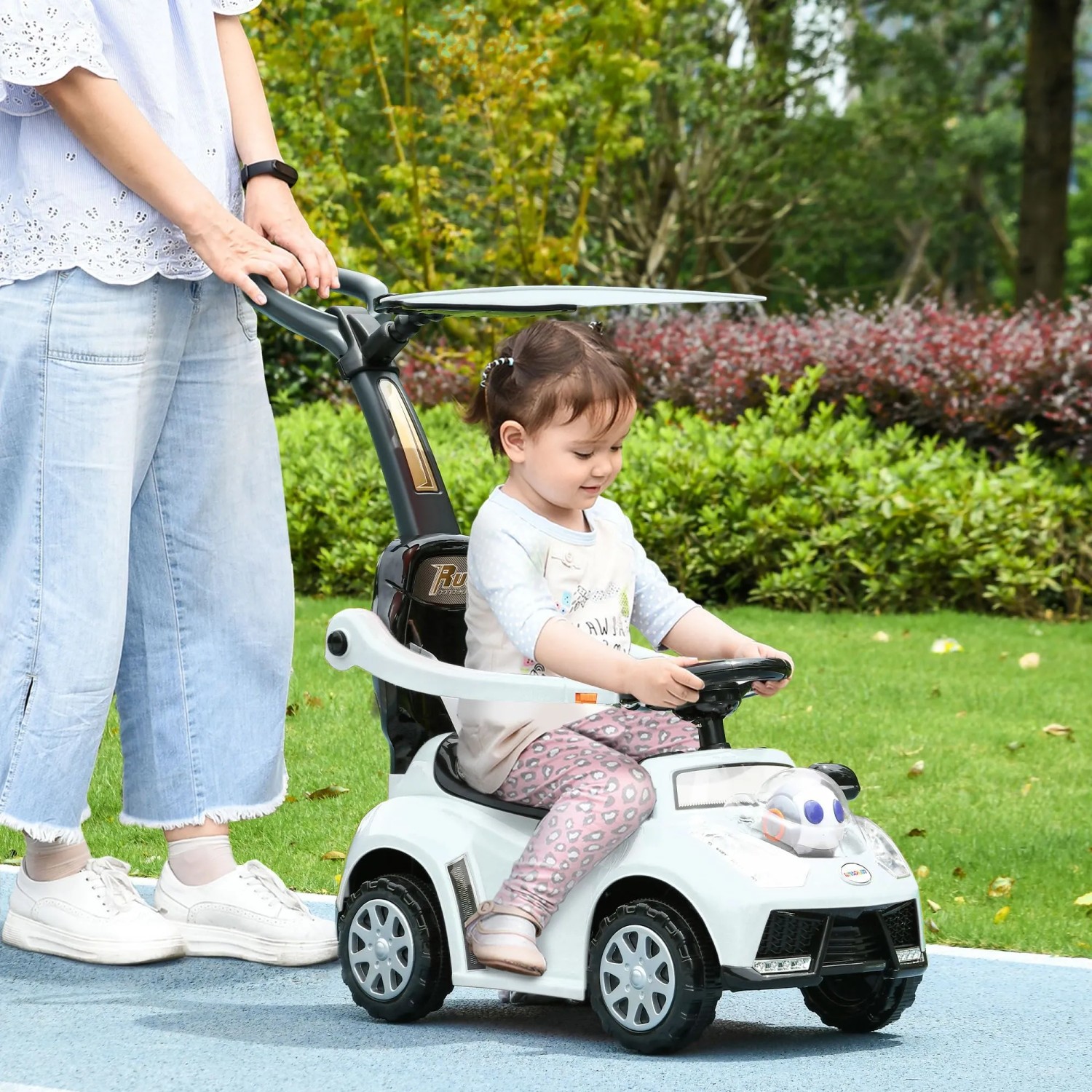 Weißes AIYAPLAY Rutschauto mit Schubstange, Rückenlehne und Sonnendach; Kind fährt, Erwachsener schiebt im Park.