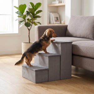Kleiner Hund auf grauer 3-Stufen Hundetreppe mit Stauraum neben Sofa. Faltbare Tiertreppe aus MDF/Polyester als Aufstiegshilfe.