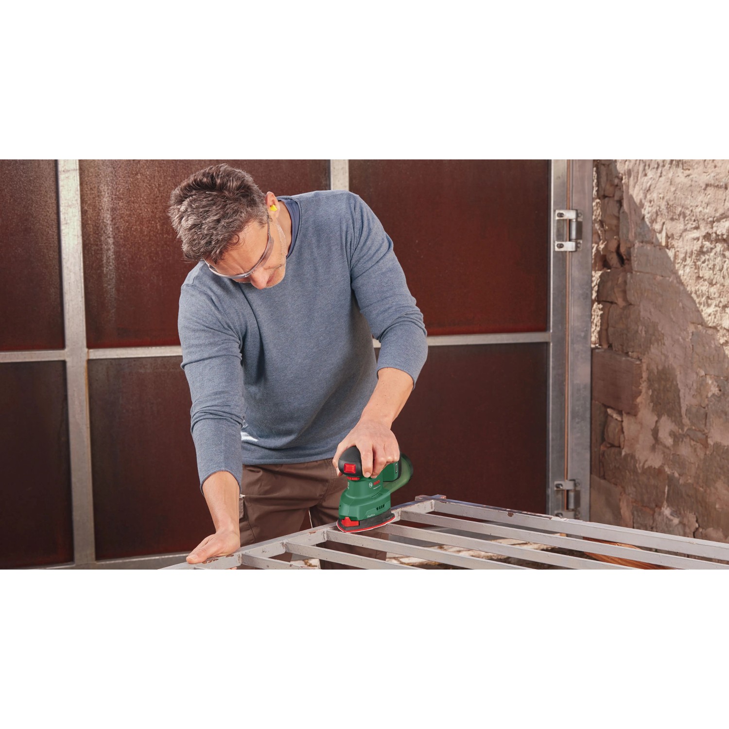 Mann schleift Metallrahmen mit Bosch Akku-Multischleifer UniversalSander 18V-10. Kompakte Schleifmaschine für präzise Arbeiten.