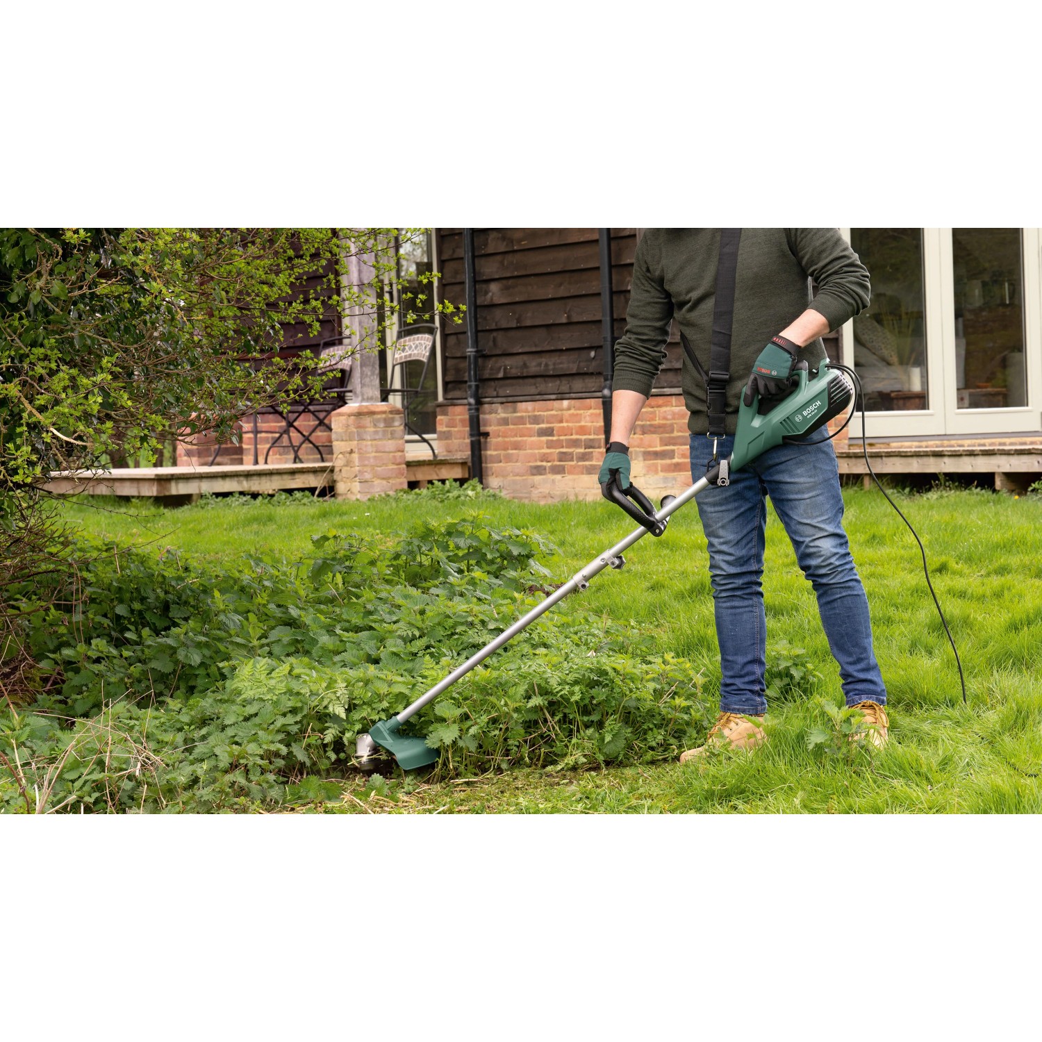 Mann trimmt Wildwuchs mit grünem Bosch Elektro-Rasentrimmer AFS 23-37 im Garten. Leistungsstarker Trimmer für Rasenpflege.