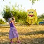 Kind schlägt auf gelbe Monster Pinata aus Pappe mit drei Augen. Bunte Partydekoration für Kindergeburtstag.