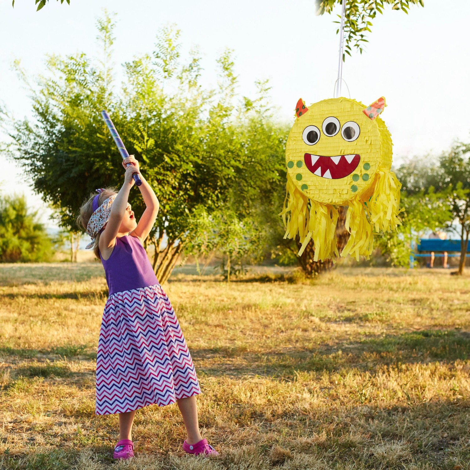 Kind schlägt auf gelbe Monster Pinata aus Pappe mit drei Augen. Bunte Partydekoration für Kindergeburtstag.