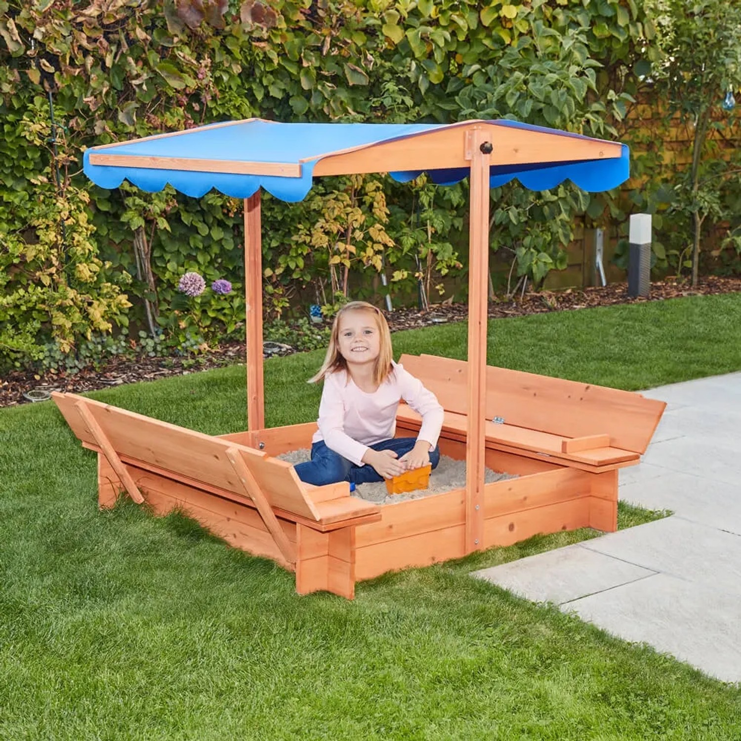 Home Deluxe Sandkasten Buddelkiste mit blauem Dach und Kind beim Spielen im Garten.