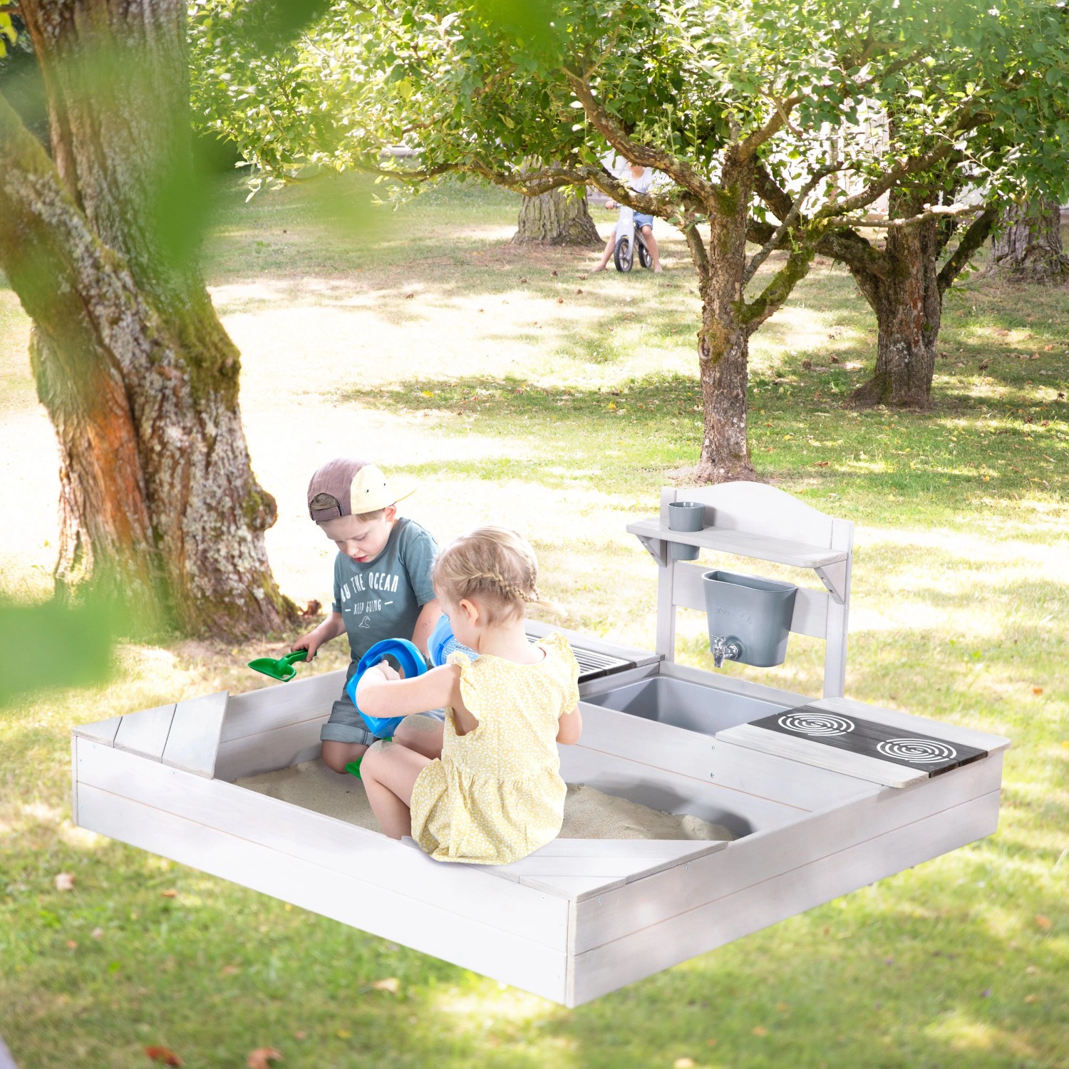 roba Sandkasten aus grau lasiertem Holz mit Matschküche, spielenden Kindern, Sitzbänken, Herdplatten und Wasserhahn.